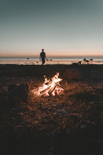 View of a person standing on the beach of Vormsi island near a glowing campfire at sunset with calm sea and rocky shoreline Laane County Estonia. View of a person standing on the beach of Vormsi island near a glowing campfire at sunset with calm sea and rocky shoreline Laane County Estonia.