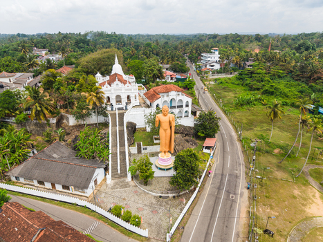 Aerial view of the white Kumarakanda Kumara Maha Viharaya temple and a large golden standing Buddha statue surrounded by lush tropical greenery Hikkaduwa Southern Province Sri Lanka. Aerial view of the white Kumarakanda Kumara Maha Viharaya temple and a large golden standing Buddha statue surrounded by lush tropical greenery Hikkaduwa Southern Province Sri Lanka.