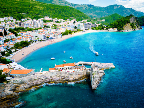Aerial view of Kastio Castle on a rocky peninsula with stone walls and red tiled roofs overlooking the turquoise Adriatic Sea sandy beach and Petrovac Budva Municipality Montenegro. Aerial view of Kastio Castle on a rocky peninsula with stone walls and red tiled roofs overlooking the turquoise Adriatic Sea sandy beach and Petrovac Budva Municipality Montenegro.