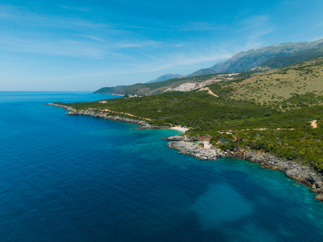 Aerial view of Aquarium Beach featuring turquoise waters and rocky cliffs along the lush green coastline in Jala Vlore County Albania. Aerial view of Aquarium Beach featuring turquoise waters and rocky cliffs along the lush green coastline in Jala Vlore County Albania.