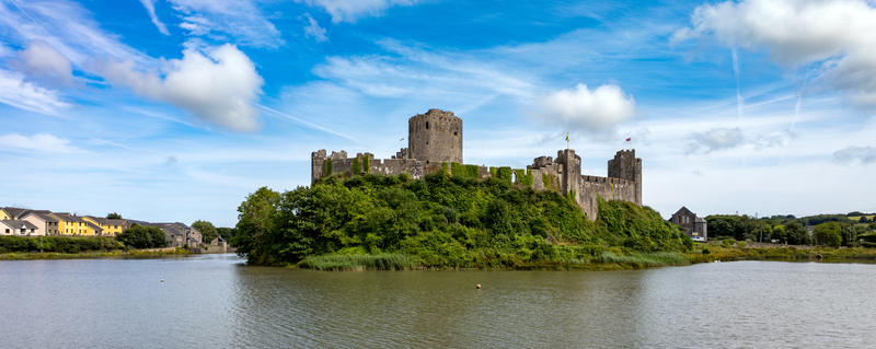 View of Pembroke Castle standing on a lush green hill overlooking the calm water under a blue sky with wispy clouds in Pembroke Wales United Kingdom.