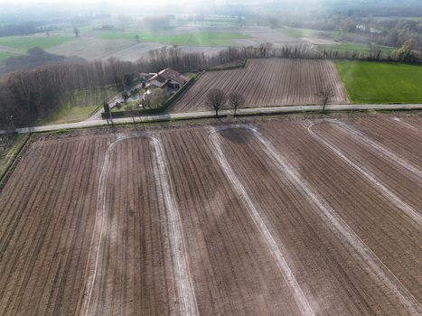 Aerial view of plowed agricultural fields with white tracks a farmhouse and a rural road under a misty sky in Borgo d Ale Piedmont Italy.