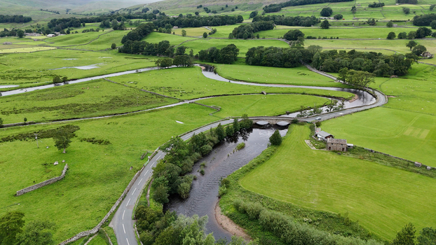 View of the winding River Ure and a stone bridge crossing a road amidst the lush green pastures and stone walls of the Yorkshire Dales in Hawes England United Kingdom. View of the winding River Ure and a stone bridge crossing a road amidst the lush green pastures and stone walls of the Yorkshire Dales in Hawes England United Kingdom.