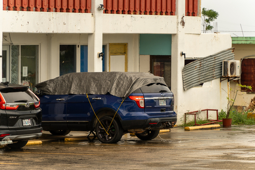 San Jose Tinian Northern Mariana Islands 05 July 2026 View of a blue SUV covered with a tarp parked in front of a building with damaged siding and an air conditioning unit. San Jose Tinian Northern Mariana Islands 05 July 2026 View of a blue SUV covered with a tarp parked in front of a building with damaged siding and an air conditioning unit.