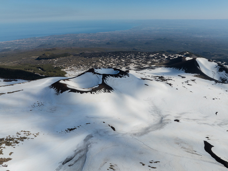 Aerial view of the snow covered volcanic craters on the Versante sud dell Etna with dark lava fields and the distant coastline above Nicolosi Sicilia Italy. Aerial view of the snow covered volcanic craters on the Versante sud dell Etna with dark lava fields and the distant coastline above Nicolosi Sicilia Italy.