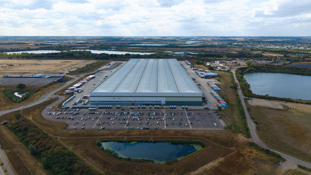 Aerial view of a large logistics hub with industrial warehouses rows of parked HGVs and a busy car park set within a landscape of lakes and fields in Bedford England United Kingdom. Aerial view of a large logistics hub with industrial warehouses rows of parked HGVs and a busy car park set within a landscape of lakes and fields in Bedford England United Kingdom.