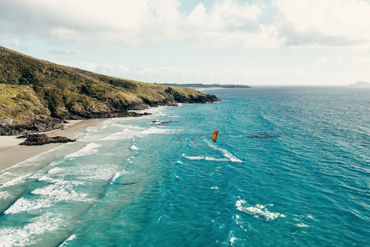 Aerial view of Puwheke Beach with a kite surfer gliding over turquoise waves along the rugged coastline of Karikari Peninsula Northland Region New Zealand.