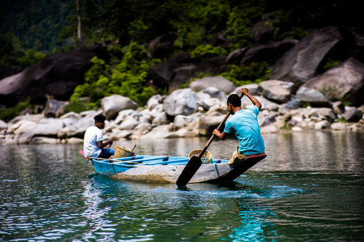 Meghalaya India 31 March 2023 View of two men in a small blue wooden boat rowing on a river with large boulders along the bank. Meghalaya India 31 March 2023 View of two men in a small blue wooden boat rowing on a river with large boulders along the bank.
