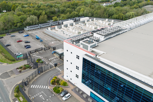 Aerial view of a modern data center facility with rooftop cooling units a secure parking area and surrounding green landscape in the United Kingdom.