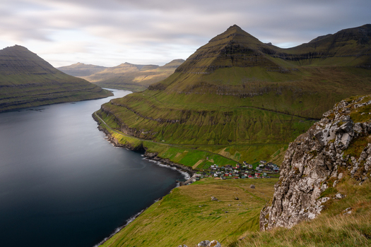 View of a small village nestled along the coast of a deep fjord surrounded by steep grass covered mountains under a soft cloudy sky in the Faroe Islands. View of a small village nestled along the coast of a deep fjord surrounded by steep grass covered mountains under a soft cloudy sky in the Faroe Islands.