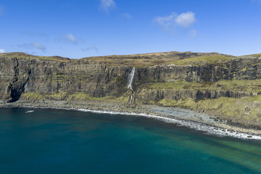 Aerial view of Talisker Bay Beach featuring a waterfall cascading down steep rocky cliffs into the turquoise sea under a clear blue sky Isle of Skye Scotland United Kingdom. Aerial view of Talisker Bay Beach featuring a waterfall cascading down steep rocky cliffs into the turquoise sea under a clear blue sky Isle of Skye Scotland United Kingdom.