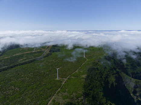 Aerial view of wind turbines situated on the lush green ridge of Bica da Cana peak above a dense layer of clouds under a clear blue sky in Funchal Madeira Portugal.