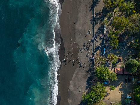 Aerial view of the Ngelarung Sea Offering Ceremony at Pura Segara Goa Lawah with people in traditional attire on the black sand beach by the turquoise ocean in Dawan Bali Indonesia.
