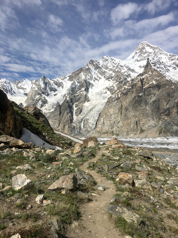 View of the rugged Hushe River Valley with a rocky trail leading towards snow capped peaks and a glacier under a partly cloudy sky Saling Gilgit Pakistan. View of the rugged Hushe River Valley with a rocky trail leading towards snow capped peaks and a glacier under a partly cloudy sky Saling Gilgit Pakistan.