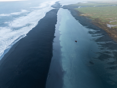 Aerial view of a small boat floating in a turquoise river channel alongside a vast black sand beach and green coastal plains in Iceland. Aerial view of a small boat floating in a turquoise river channel alongside a vast black sand beach and green coastal plains in Iceland.
