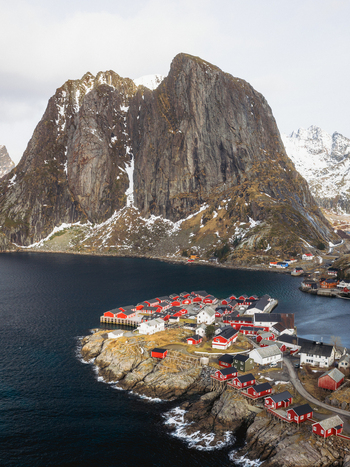 Aerial view of red cabins contrasting with the dark waters and the rugged snow dusted mountain peaks of Sakrisøya Nordland Norway. Aerial view of red cabins contrasting with the dark waters and the rugged snow dusted mountain peaks of Sakrisøya Nordland Norway.