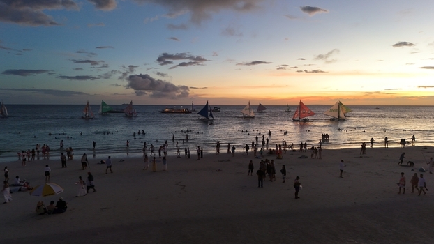 Malay Philippines 02 May 2026 Aerial view of Boracay Island featuring crowds of people on the white sand beach and traditional sailboats on the calm sea during a vibrant sunset.