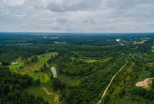 Aerial view of a lush green forest landscape with a small pond and a winding dirt path under a cloudy sky in Latvia. Aerial view of a lush green forest landscape with a small pond and a winding dirt path under a cloudy sky in Latvia.