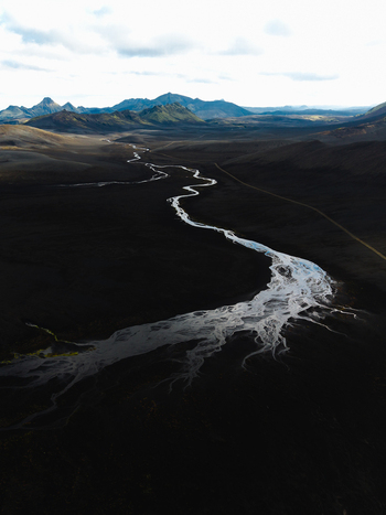 Aerial view of a winding river snaking through a vast dark volcanic landscape contrasting with distant green hills under a cloudy sky Iceland. Aerial view of a winding river snaking through a vast dark volcanic landscape contrasting with distant green hills under a cloudy sky Iceland.