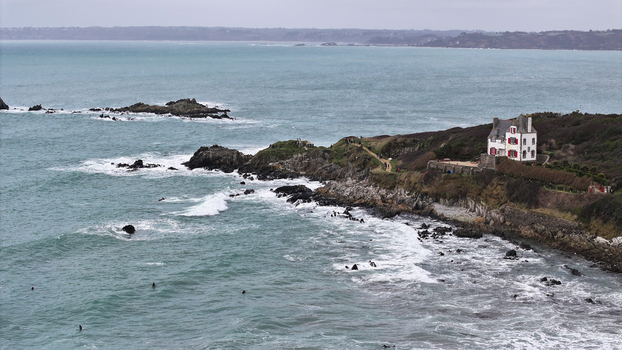 Aerial view of the rugged coastline of Locquirec featuring a white house with red shutters on a cliff overlooking the turquoise sea and surfers in the waves Locquirec Brittany France.