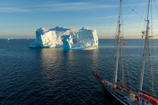 Aerial view of a majestic arch iceberg floating in the deep blue sea with a sailing ship in the foreground in Greenland. Aerial view of a majestic arch iceberg floating in the deep blue sea with a sailing ship in the foreground in Greenland.