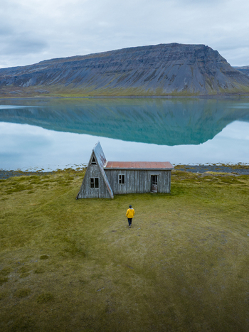 Aerial view of a solitary figure in a yellow jacket standing before a weathered building with a distinctive A frame roof framed by tranquil waters and rugged mountains Westfjords Iceland. Aerial view of a solitary figure in a yellow jacket standing before a weathered building with a distinctive A frame roof framed by tranquil waters and rugged mountains Westfjords Iceland.