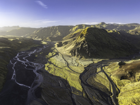 Aerial view of the braided river channels winding through mossy volcanic hills and rugged mountain peaks under a clear sky in Katla Geopark Skaftarhreppur Iceland.