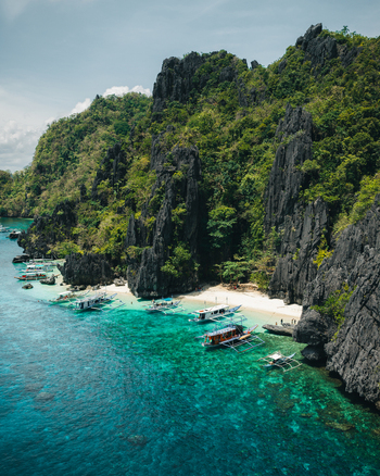 Aerial view of limestone cliffs rising from turquoise waters with traditional outrigger boats moored along a sandy beach in El Nido Mimaropa Philippines. Aerial view of limestone cliffs rising from turquoise waters with traditional outrigger boats moored along a sandy beach in El Nido Mimaropa Philippines.