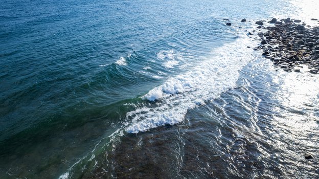 Aerial view of ocean waves crashing against a rocky shoreline with clear blue water and sunlight reflecting on the surface Bajawa Nusa Tenggara Timur Indonesia. Aerial view of ocean waves crashing against a rocky shoreline with clear blue water and sunlight reflecting on the surface Bajawa Nusa Tenggara Timur Indonesia.