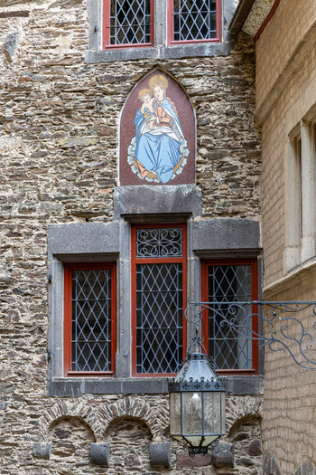 View of the medieval stone facade of Eltz Castle featuring a religious mosaic of the Virgin Mary and Child above red framed windows and a wrought iron lantern in Wierschem Rhineland Palatinate Germany.
