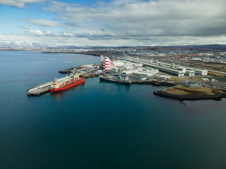 Hafnarfjordur Iceland 17 April 2026 Aerial view of the industrial port with a large red cargo ship docked near warehouses and a striped silo under a cloudy sky. Hafnarfjordur Iceland 17 April 2026 Aerial view of the industrial port with a large red cargo ship docked near warehouses and a striped silo under a cloudy sky.