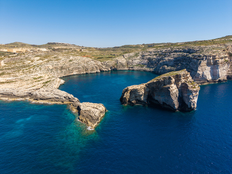 Aerial view of Dwejra Bay featuring rugged limestone cliffs and deep blue Mediterranean waters under a clear sky Gozo Region Malta. Aerial view of Dwejra Bay featuring rugged limestone cliffs and deep blue Mediterranean waters under a clear sky Gozo Region Malta.