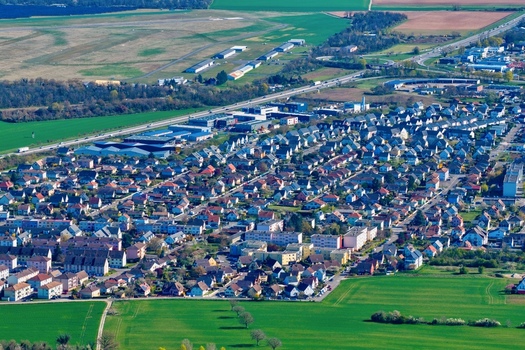 Aerial view of tightly packed houses bask in the sunlight contrasting with the surrounding green fields and distant industrial buildings Sausheim Grand Est France.