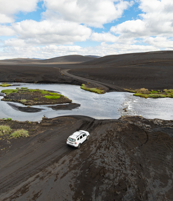 Aerial view of a white SUV driving through a shallow river crossing in the vast black volcanic landscape of Iceland. Aerial view of a white SUV driving through a shallow river crossing in the vast black volcanic landscape of Iceland.