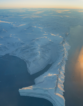 Aerial view of snow covered mountain ranges and icy fjords illuminated by the soft golden light of sunset Greenland. Aerial view of snow covered mountain ranges and icy fjords illuminated by the soft golden light of sunset Greenland.