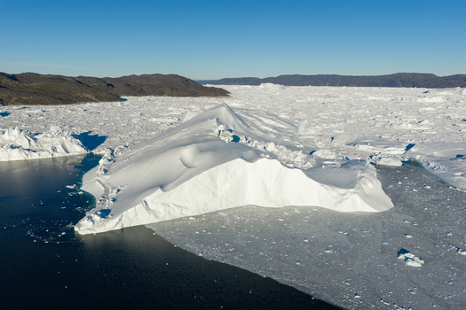 Aerial view of the Ilulissat glacier featuring a massive iceberg floating in the dark Arctic waters surrounded by extensive sea ice and rocky coastlines under a clear blue sky in Greenland. Aerial view of the Ilulissat glacier featuring a massive iceberg floating in the dark Arctic waters surrounded by extensive sea ice and rocky coastlines under a clear blue sky in Greenland.