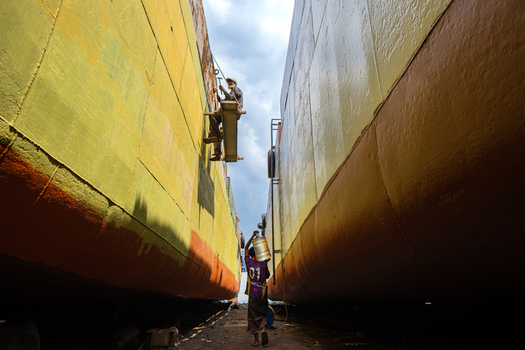 Keraniganj Bangladesh 24 September 2025 View of Shipyard Dhaka with workers repairing large ship hulls painted in yellow and orange under a cloudy sky.