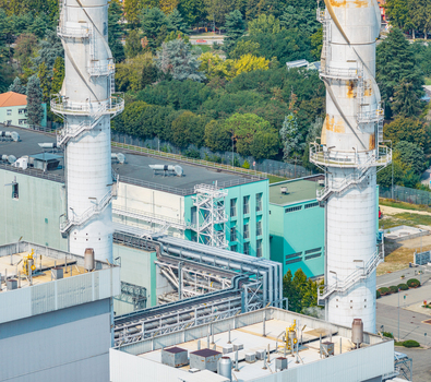Aerial view of the Chivasso combined cycle power plant featuring tall exhaust stacks industrial pipelines and complex machinery set against a backdrop of green trees in Italy.