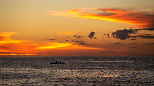 View of Bantayan Island at sunset with a small traditional fishing boat silhouetted against the vibrant orange and yellow sky over the calm sea in Cebu Central Visayas Philippines. View of Bantayan Island at sunset with a small traditional fishing boat silhouetted against the vibrant orange and yellow sky over the calm sea in Cebu Central Visayas Philippines.