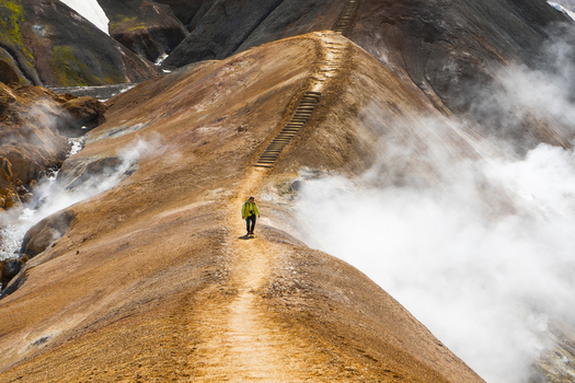 View of a hiker in a yellow jacket walking along a dirt path with wooden stairs in the geothermal landscape of Kerlingarfjoll Hrunamannahreppur Iceland.