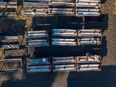 Aerial view of a traditional natural salt maker with wooden troughs filled with salt crystals and a worker harvesting on dark volcanic sand in Kecamatan Dawan Bali Indonesia. Aerial view of a traditional natural salt maker with wooden troughs filled with salt crystals and a worker harvesting on dark volcanic sand in Kecamatan Dawan Bali Indonesia.