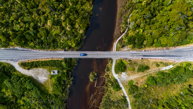 Aerial view of a single blue car crossing a bridge over the dark winding river framed by vibrant green forest Ship Creek Haast West Coast Region New Zealand. Aerial view of a single blue car crossing a bridge over the dark winding river framed by vibrant green forest Ship Creek Haast West Coast Region New Zealand.