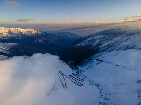 Aerial view of the winding Babusar Pass road snaking through snow covered mountain peaks under a soft sunset sky with thick clouds over the landscape Babusar KPK Pakistan. Aerial view of the winding Babusar Pass road snaking through snow covered mountain peaks under a soft sunset sky with thick clouds over the landscape Babusar KPK Pakistan.