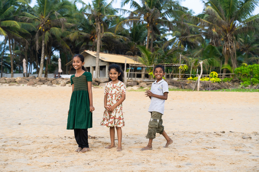 Sri Lanka 20 January 2026 View of three smiling children standing on a sandy beach with lush palm trees and a small coastal building in the background.