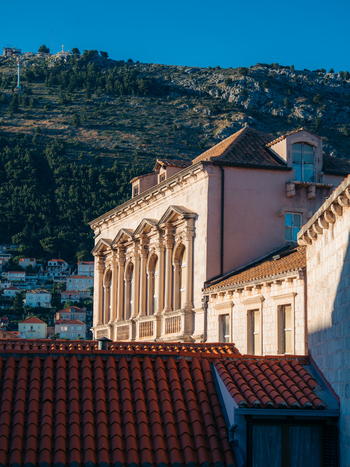 View of historic stone buildings with red tile roofs and an ornate facade against a backdrop of a steep green hill in Dubrovnik Dubrovnik Neretva County Croatia. View of historic stone buildings with red tile roofs and an ornate facade against a backdrop of a steep green hill in Dubrovnik Dubrovnik Neretva County Croatia.