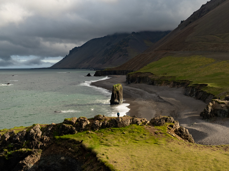 Aerial view of the dramatic coastline at Stapavik with a person on the rocky cliff overlooking the black sand beach sea waves and rugged mountains in Njardhvik Mulathing Iceland. Aerial view of the dramatic coastline at Stapavik with a person on the rocky cliff overlooking the black sand beach sea waves and rugged mountains in Njardhvik Mulathing Iceland.