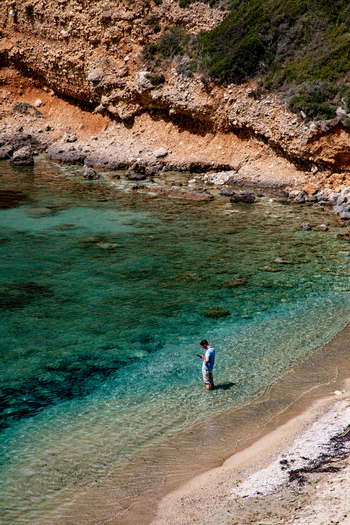 Aerial view of a man standing in the shallow turquoise water of a secluded beach with rocky cliffs in the background in Corfu Ionian Islands Greece. Aerial view of a man standing in the shallow turquoise water of a secluded beach with rocky cliffs in the background in Corfu Ionian Islands Greece.