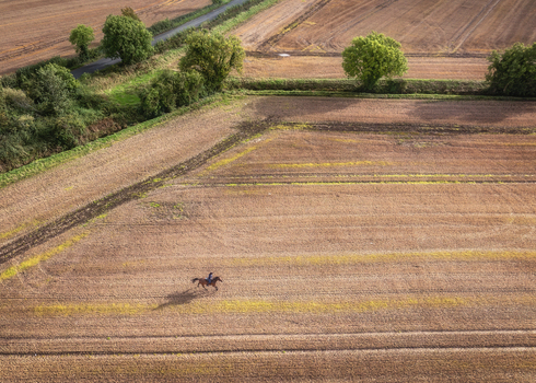 Aerial view of a person riding a horse across a large harvested agricultural field with green trees and a rural road in Oldtown County Dublin Ireland.