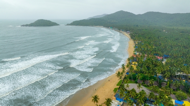 Aerial view of Palolem Beach with its crescent shaped coastline lush palm trees rolling waves and distant hills under an overcast sky in Canacona Goa India.