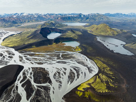 Aerial view of glacial rivers winding through volcanic landscapes dark lava fields contrasting with vibrant green moss and distant snow capped mountains Highlands Iceland.